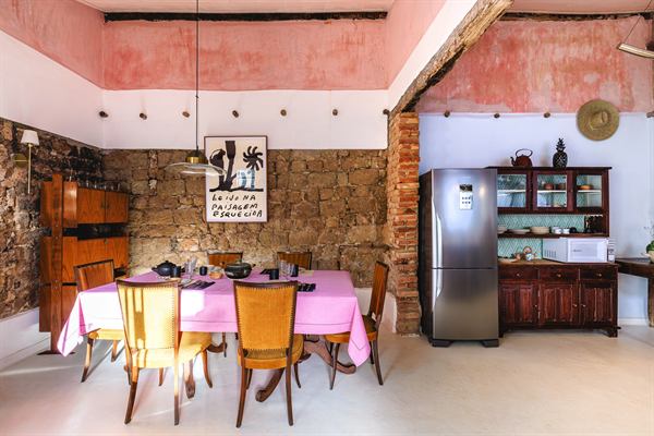 Cozinha com mesa posta de toalha rosa, parede de pedra e cristaleira antiga