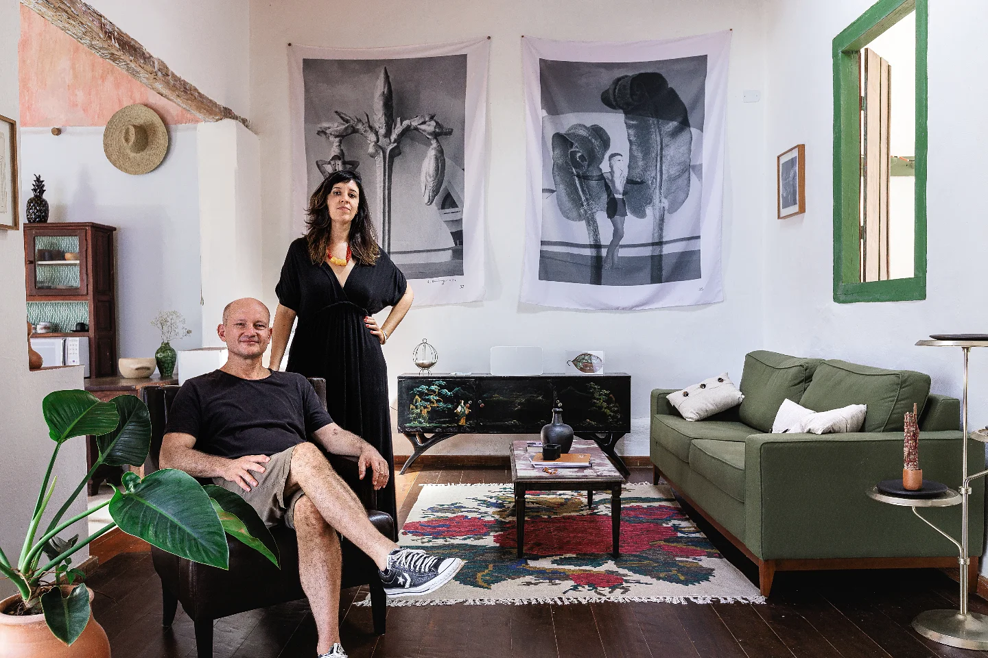 Juliana Freire, de vestido preto, em pé ao lado de Christophe Van Hamme, sentado numa poltrona, na sala da Casa Tempo, com fotografias em tecido na parede e um sofá verde