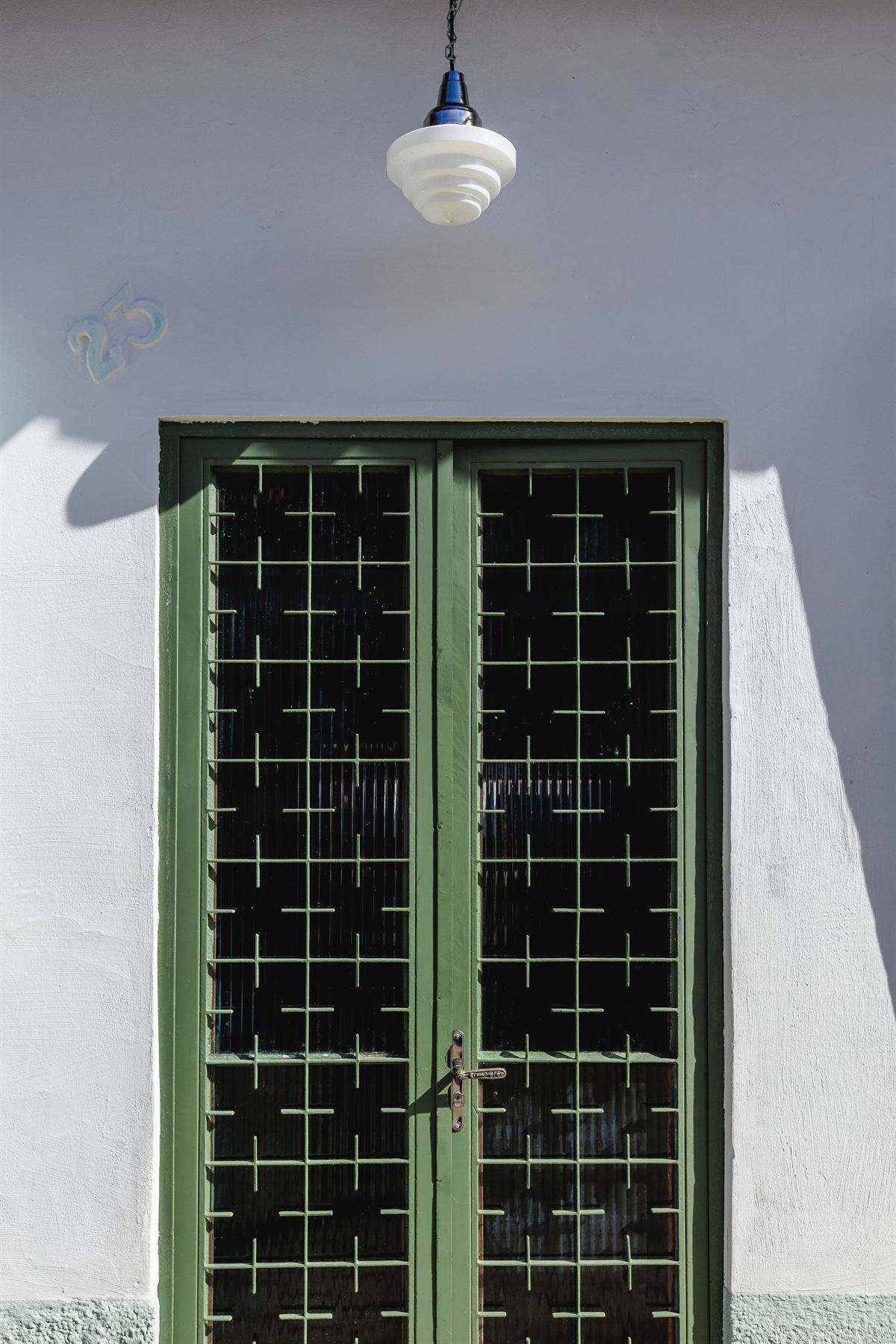Porta dupla verde com grade, em parede branca sob luminária pendente