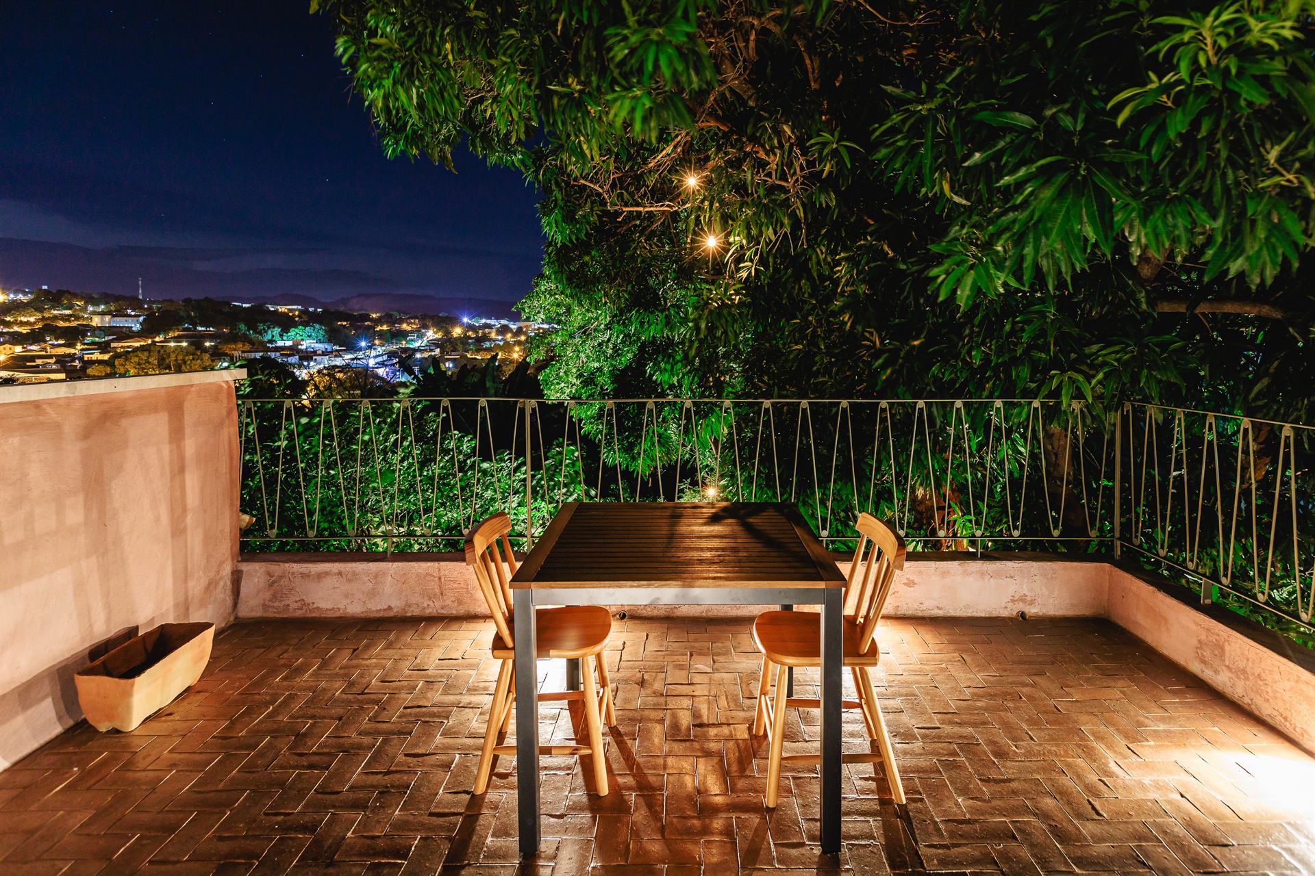 Varanda de piso de tijolos com mesa e cadeiras à noite, cercada de árvores, com as luzes da cidade ao fundo