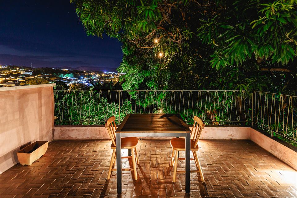 Terraço de piso de tijolos com mesa e cadeiras à noite, com as luzes da cidade ao fundo